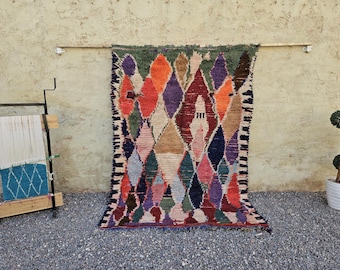 May include: A rectangular hand-woven rug featuring a repeating diamond pattern in vibrant colors including orange, purple, red, blue, green, and tan. The rug has a thick, textured pile and a dark, fringed border with a black and white striped edge.
