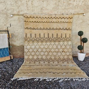 Könnte beinhalten: Beige und brauner Teppich mit geometrischem Muster, präsentiert vor einer strukturierten Wand. Der Teppich hat einen Fransenbesatz am unteren Rand. Ein Webstuhl und ein Topfbaum sind ebenfalls im Bild.