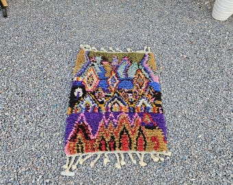 May include: A rectangular hand-knotted rug featuring a high-pile texture with a vibrant, multi-colored geometric pattern. The design includes shades of purple, blue, yellow, pink, and black, finished with cream-colored fringe on the top and bottom edges.