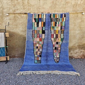Könnte beinhalten: Ein blauer Teppich mit geometrischem Design, der an einer strukturierten Wand hängt. Der Teppich hat zwei große, farbenfrohe, abstrakte Formen mit Fransen am unteren Rand. Ein kleiner Webstuhl und eine Topiarkugel im Topf sind im Hintergrund.