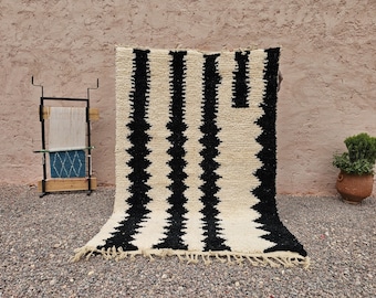 May include: A large, cream-colored rug with bold black vertical stripes and a fringed edge. The rug is displayed outdoors, leaning against a textured wall. A small plant in a terracotta pot is visible.