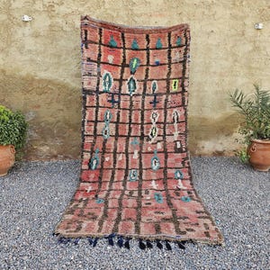Puede incluir: Una alfombra rectangular tejida a mano con un patrón geométrico en tonos de rojo, negro y verde azulado. La alfombra presenta un diseño similar a una cuadrícula con formas abstractas y un borde con flecos. La alfombra se exhibe al aire libre, apoyada contra una pared texturizada.