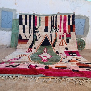 May include: A large, colorful Moroccan rug with geometric patterns in shades of red, pink, green, black, and white. The rug is hanging on a wall and has a fringe border.