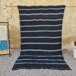 May include: A black rug with horizontal white and blue stripes hangs against a beige wall. The rug's woven texture is visible. A wooden loom and a potted topiary are in the background, adding to the rustic aesthetic.