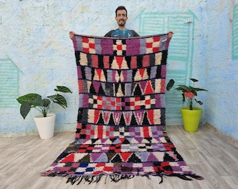 May include: A colorful Moroccan rug with a geometric pattern in shades of red, purple, blue, and white. The rug is hand-woven and has a fringe border.