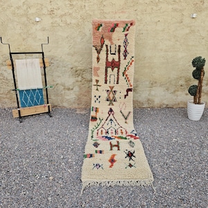 Peut inclure: Un long tapis de couloir étroit avec une base de couleur crème ornée de divers motifs géométriques en rouge, vert, bleu et marron. Le tapis présente une bordure à franges à la base et affiche une série de symboles et de formes abstraites.