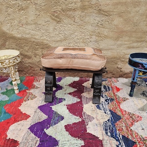 Peut inclure: Trois tabourets décoratifs en bois. L'un est blanc avec des détails dorés, un autre a un coussin en cuir beige et le troisième est bleu avec des motifs colorés. Ils sont présentés sur un tapis aux motifs vibrants.