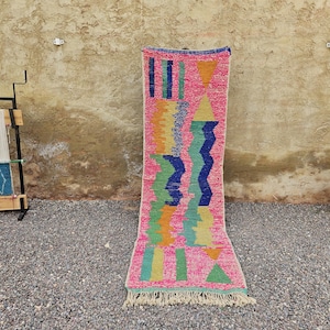 May include: A colourful, rectangular rug with a pink base and geometric patterns. The design features blue, green, orange, and yellow shapes. The rug has a fringed edge and is displayed outdoors.