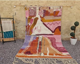 May include: A rectangular hand-woven wool rug featuring an abstract geometric pattern in shades of pink, orange, mustard yellow, purple, and cream. The rug has a thick fringe along the bottom edge and a textured pile with varied color blocks.