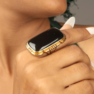 May include: A gold-toned ring featuring a large, rectangular black stone. The ring has a detailed, geometric design around the stone and on the band. The ring is worn on a finger, showcasing its bold and elegant style.