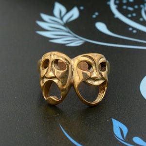 May include: A gold-colored ring featuring two stylized faces, one with a smiling expression and the other with a crying expression. The ring is a classic representation of comedy and tragedy.