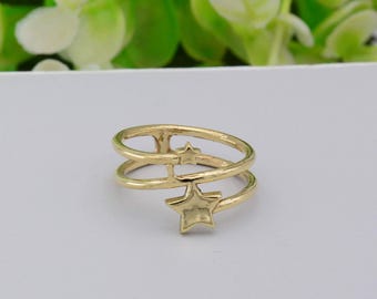 May include: A gold ring with a star design. The ring has two bands connected by a star-shaped charm and a smaller star. The ring is set against a white surface with green foliage in the background.