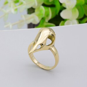 Könnte beinhalten: Ein goldfarbener Ring mit einem Vogelschädel-Design. Der Ring hat einen detaillierten, durchbrochenen Schädel mit Augen- und Nasenöffnungen. Der Ring ist glatt, was einen Kontrast zum Schädel bildet. Ein einzigartiges Schmuckstück.