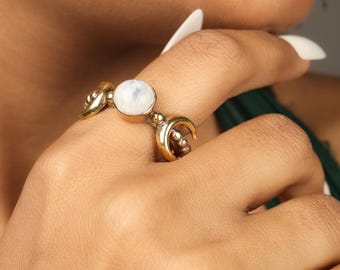 May include: A gold-coloured ring with a central, round, white gemstone, and crescent moon and decorative elements. The ring is worn on a finger, highlighting its intricate design and celestial theme. The ring's design is detailed and eye-catching.