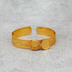 May include: A gold-toned bangle bracelet with an ornate floral design. The bracelet has two round, open-backed settings for adding charms or stones.