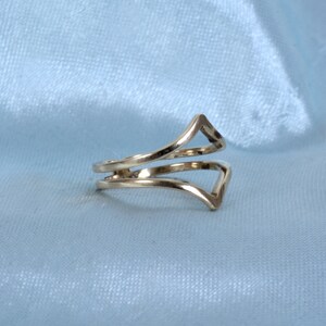 May include: A gold-colored ring with a unique design. The ring features two bands that wrap around the finger, with a V-shaped element at the top. The ring is displayed on a light blue background.