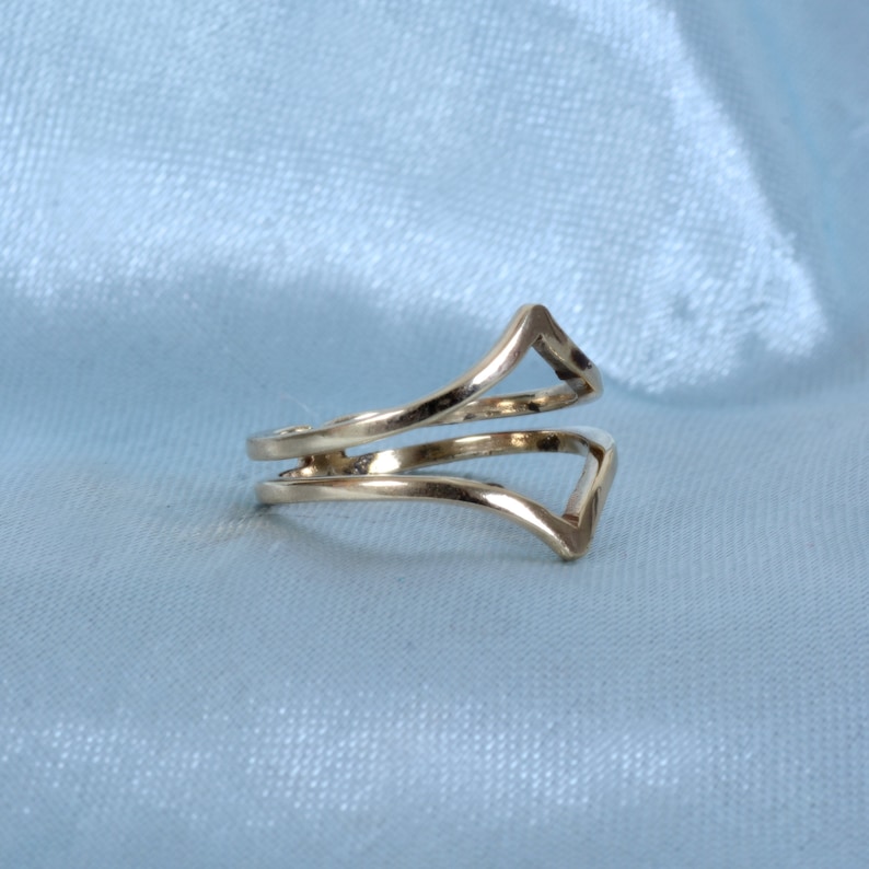 May include: A gold-colored ring with a unique design. The ring features two bands that wrap around the finger, with a V-shaped element at the top. The ring is displayed on a light blue background.