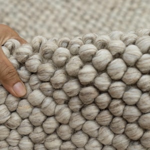 Può includere: Primo piano di un tappeto beige chiaro strutturato con una superficie nodosa a forma di bolla. Il tappeto viene sollevato, rivelando la sua costruzione e il retro tessuto. La trama generale è morbida e invitante, adatta all'arredamento.