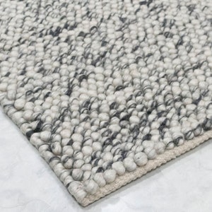 Puede incluir: Primer plano de una alfombra de lana blanca y gris con una superficie texturizada y abultada. La alfombra tiene un sutil patrón de hilo negro y blanco.