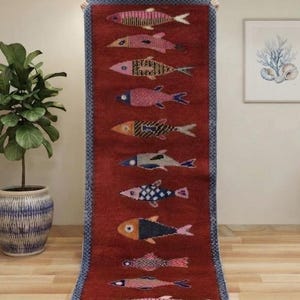 May include: A long, rectangular runner rug with a deep red background featuring a vertical row of thirteen stylized fish in various colors and patterns. The rug has a blue patterned border and white fringe at the bottom edge.
