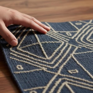 Puede incluir: Una alfombra azul con un patrón geométrico crema. El diseño de la alfombra presenta líneas y formas, creando una estética moderna. La alfombra es rectangular y tiene una superficie texturizada. Está colocada sobre un suelo de madera.