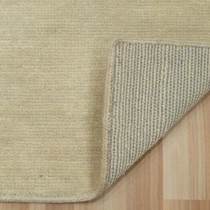 May include: A rectangular area rug with a solid cream-colored, textured pile surface. One corner of the rug is folded back to reveal a light gray, grid-patterned backing material with a visible stitched edge along the perimeter of the rug.