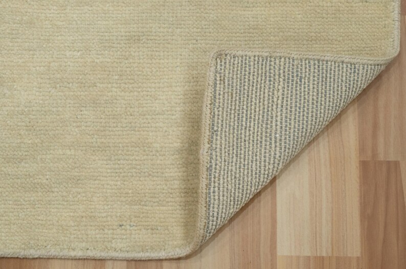 May include: A rectangular area rug with a solid cream-colored, textured pile surface. One corner of the rug is folded back to reveal a light gray, grid-patterned backing material with a visible stitched edge along the perimeter of the rug.