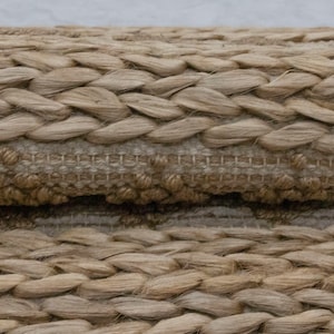 May include: Close-up of a woven rug made of natural fibers. The rug has a braided texture and a light brown color.