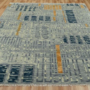 Puede incluir: Alfombra rectangular con un patrón geométrico en tonos grises, azules y dorados. La alfombra tiene un aspecto texturizado y flecos en los bordes. El diseño presenta formas rectangulares y lineales.