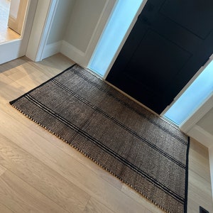May include: A black and brown striped jute rug with a fringed edge. The rug is placed on a light wood floor in front of a black door with a frosted glass panel.