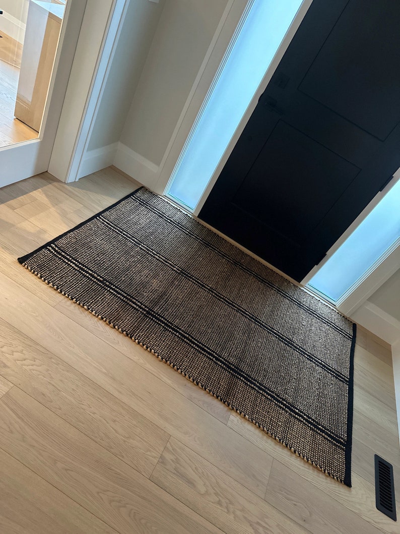 May include: A black and brown striped jute rug with a fringed edge. The rug is placed on a light wood floor in front of a black door with a frosted glass panel.
