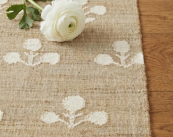 May include: A rectangular woven runner featuring a tan base with a repeating white floral pattern. A single white ranunculus flower with green leaves is placed on the surface of the runner, which has a textured, natural fiber appearance.