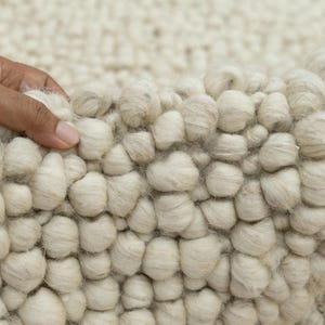May include: Close-up of a white, fluffy rug with a textured, bumpy surface. The rug is made of wool and has a soft, plush feel.