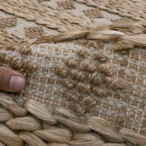 May include: A close-up of a natural fiber rug with a woven pattern. The rug is made of jute and has a light brown color. The rug is textured and has a hand-woven look.