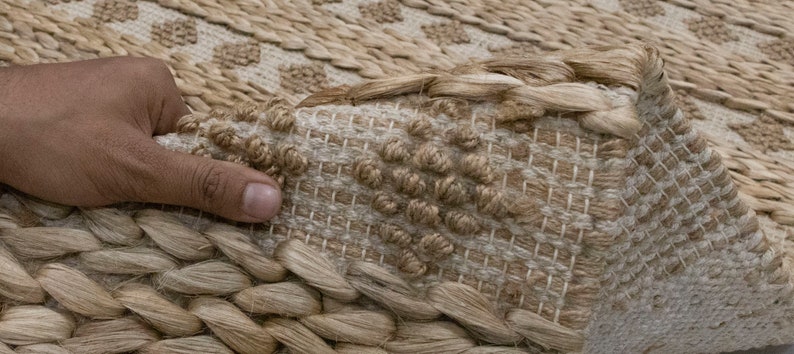 May include: A close-up of a natural fiber rug with a woven pattern. The rug is made of jute and has a light brown color. The rug is textured and has a hand-woven look.