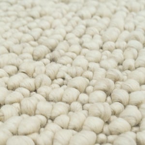 May include: Close-up of a textured, off-white rug. The rug's surface is covered in tightly packed, rounded tufts, creating a soft, tactile appearance. The neutral color and dense texture suggest a cozy, inviting feel, suitable for various interior design styles.