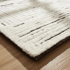 May include: A close-up of a white rug with a geometric pattern of dark brown lines. The rug has a thick, textured pile and a dark brown border. The rug is placed on a light brown wooden floor.