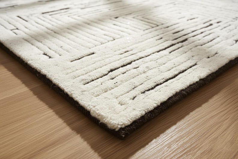 May include: A close-up of a white rug with a geometric pattern of dark brown lines. The rug has a thick, textured pile and a dark brown border. The rug is placed on a light brown wooden floor.
