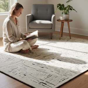May include: A cream-colored rug with a geometric black line design. The rug is rectangular and has a black border. The rug is placed on a hardwood floor in a well-lit room. A person is sitting on the rug reading a book.