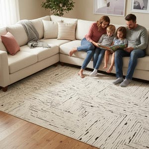 May include: A cream-colored sectional sofa with throw pillows and a gray blanket. A family of four is sitting on the sofa, reading a book. A patterned rug covers the floor, and a side table with a lamp is visible.