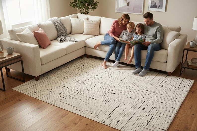 May include: A cream-colored sectional sofa with throw pillows and a gray blanket. A family of four is sitting on the sofa, reading a book. A patterned rug covers the floor, and a side table with a lamp is visible.