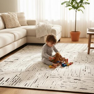 May include: A neutral-toned area rug with a geometric pattern of black lines. A child in gray pajamas sits on the rug playing with a teddy bear and wooden blocks. A beige sectional sofa and a potted plant are in the background.