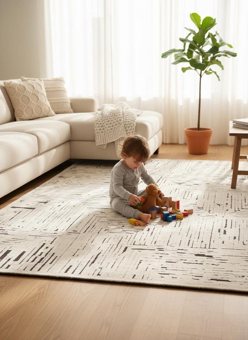 May include: A neutral-toned area rug with a geometric pattern of black lines. A child in gray pajamas sits on the rug playing with a teddy bear and wooden blocks. A beige sectional sofa and a potted plant are in the background.