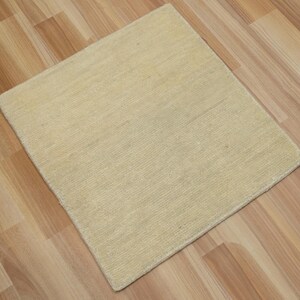 May include: A square area rug in a solid light beige color with a textured, woven surface. The rug features a consistent, dense pile construction with visible horizontal fibers throughout, creating a uniform and neutral appearance for floor coverage.
