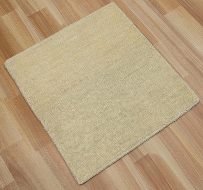 May include: A square area rug in a solid light beige color with a textured, woven surface. The rug features a consistent, dense pile construction with visible horizontal fibers throughout, creating a uniform and neutral appearance for floor coverage.