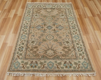 May include: A rectangular beige rug with an intricate floral pattern and a border featuring blue and brown floral motifs. The rug has a fringe on the short ends and is placed on a wooden floor. The rug's design includes shades of beige, brown, and blue.