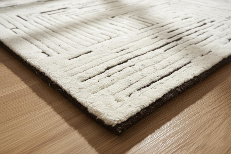 May include: A close-up of a white rug with a geometric pattern of dark brown lines. The rug has a thick, textured pile and a dark brown border. The rug is placed on a light brown wooden floor.