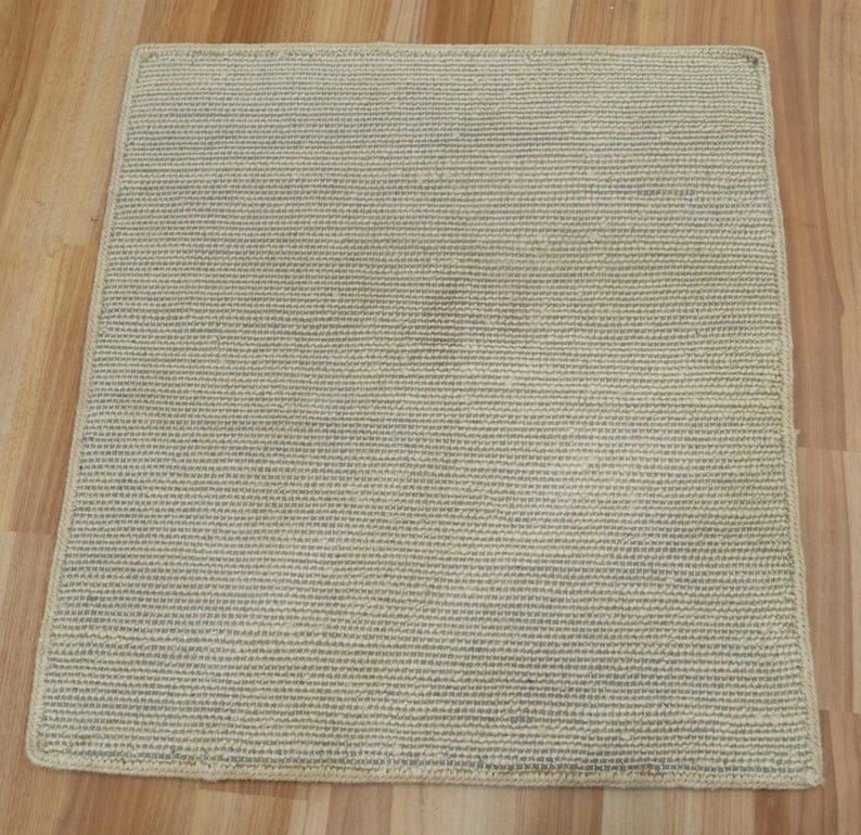 May include: A square, light beige rug featuring a dense, uniform woven texture. The rug has a consistent grid-like pattern across its entire surface, with clean, straight edges and a neutral color tone that provides a simple, flat appearance.