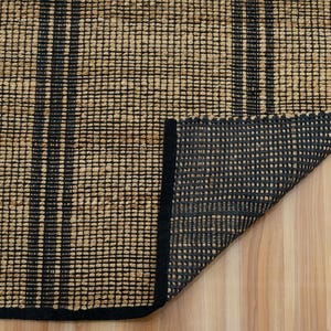 May include: A close-up of a woven rug with black stripes. The rug is made of natural fibers and has a black border. The rug is folded in half, showing the underside.