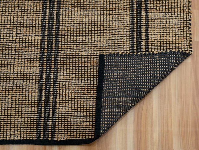 May include: A close-up of a woven rug with black stripes. The rug is made of natural fibers and has a black border. The rug is folded in half, showing the underside.